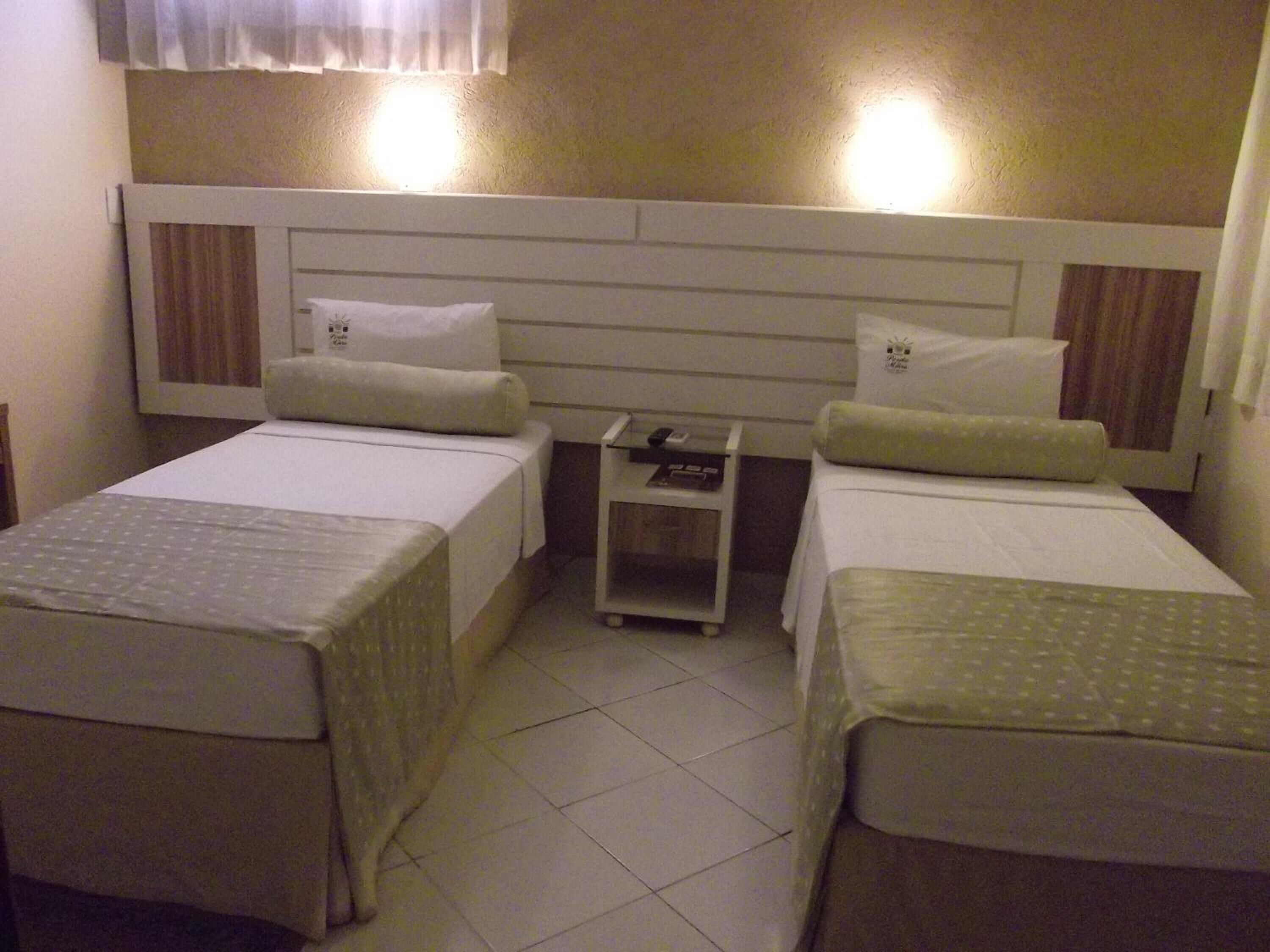Quarto de hotel com duas camas de solteiro, cada uma com travesseiro e uma pequena mesa de cabeceira.