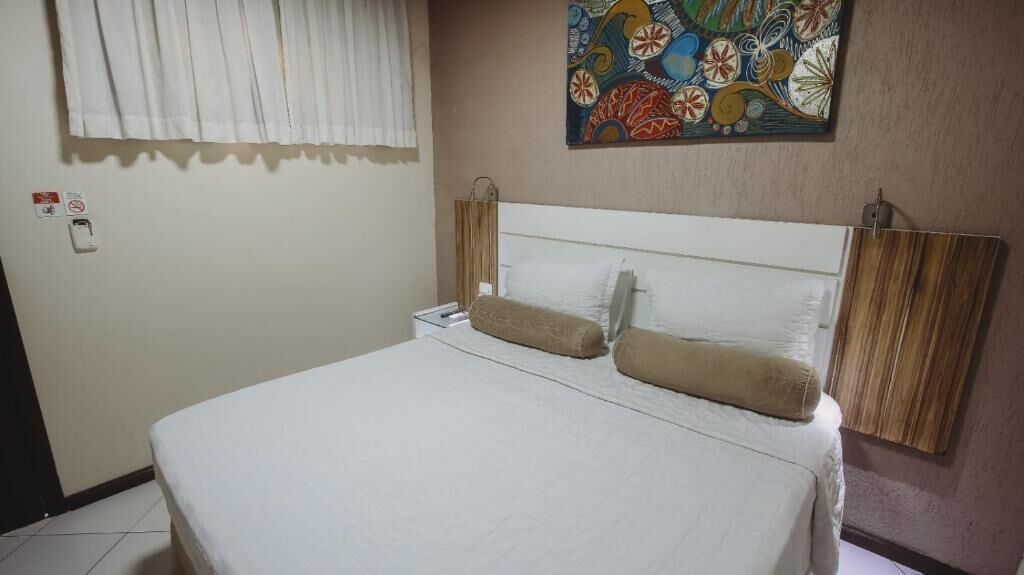Une chambre d’hôtel avec un lit, une table de chevet, un tableau au mur et une fenêtre avec des stores.