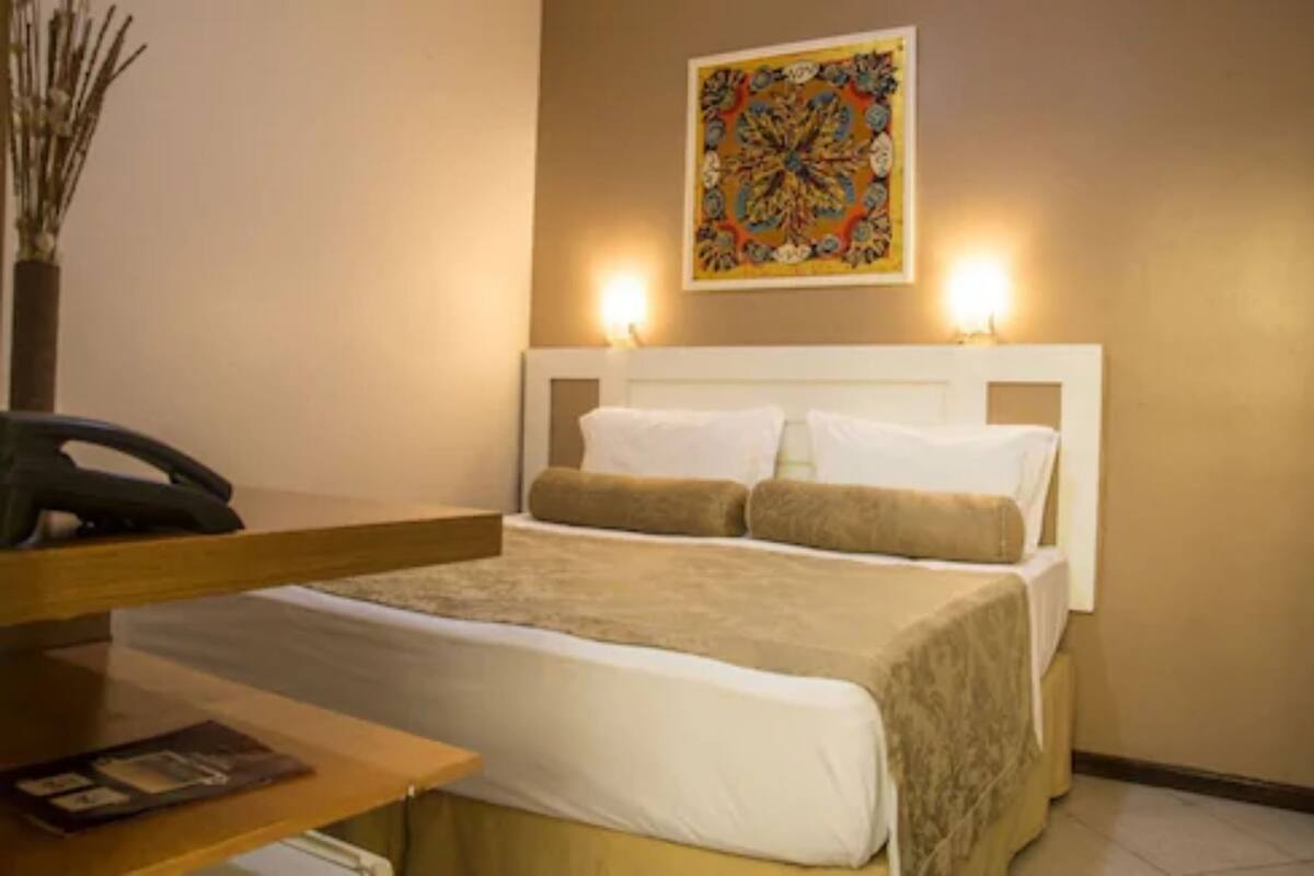 Quarto de hotel com cama, escrivaninha, cadeira e um objeto decorativo na parede.