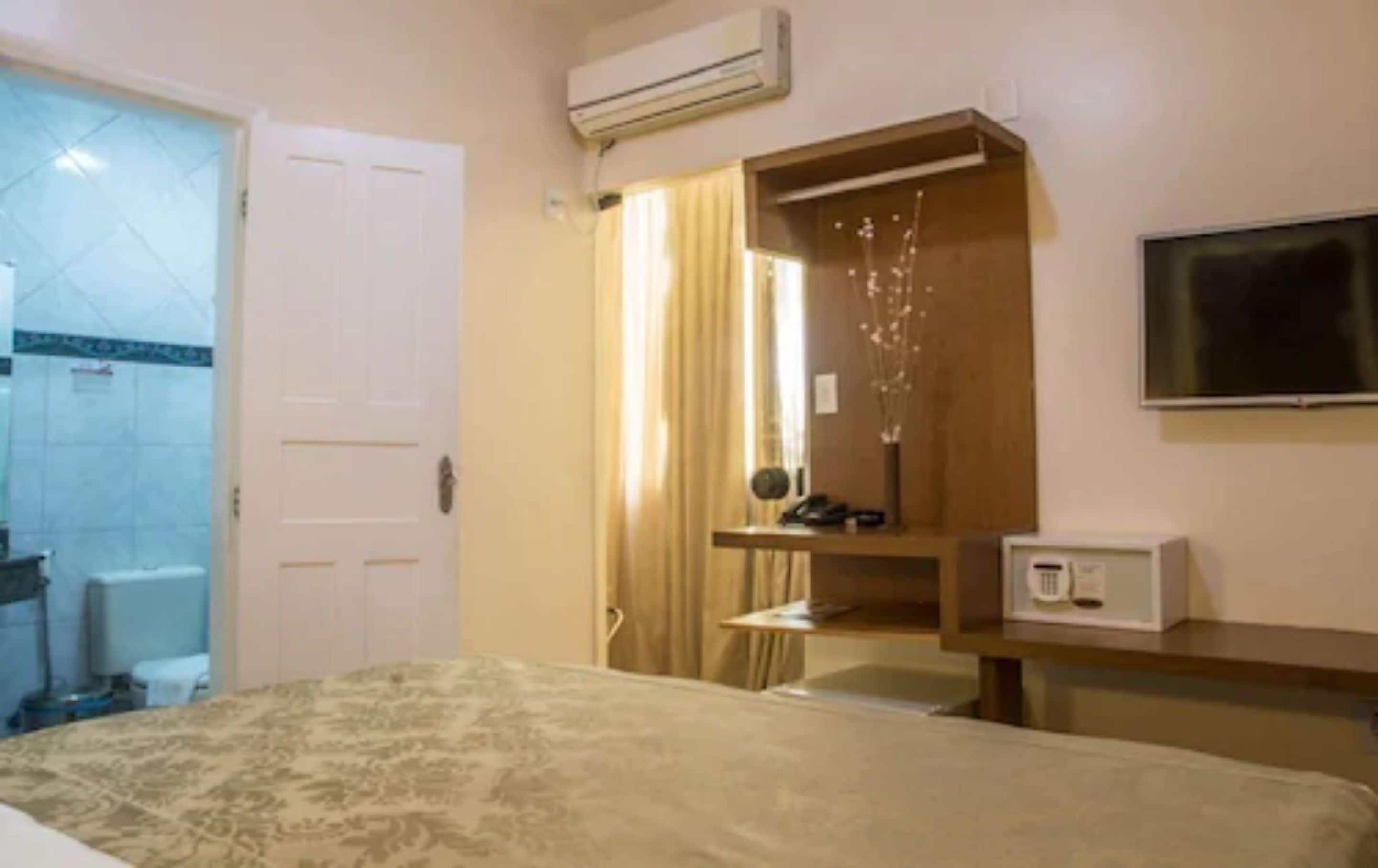 Quarto de hotel com cama, secretária, ar condicionado, televisão e casa de banho visível através de uma porta aberta.