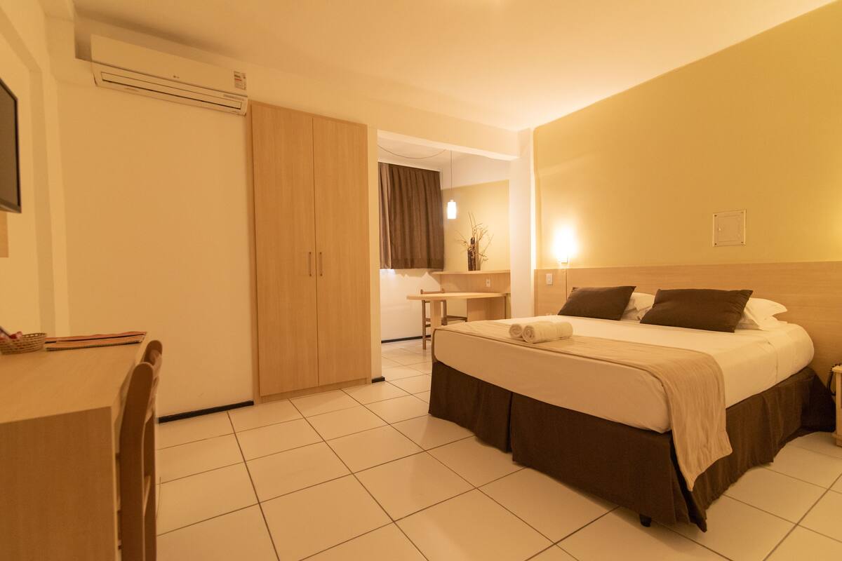 Habitación de hotel con cama, escritorio y silla. Incluye un armario y una mesita con un florero.