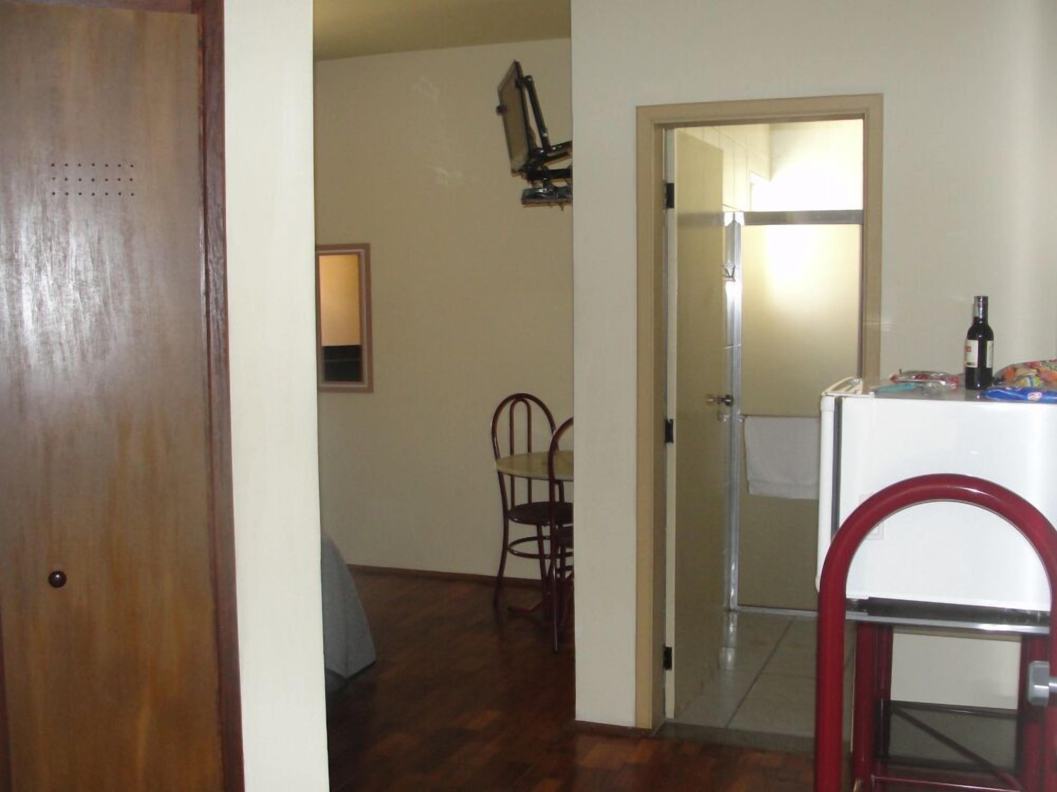 Una habitación con puerta de madera, una pequeña mesa con una silla, una pared blanca y una puerta que da a otra habitación.