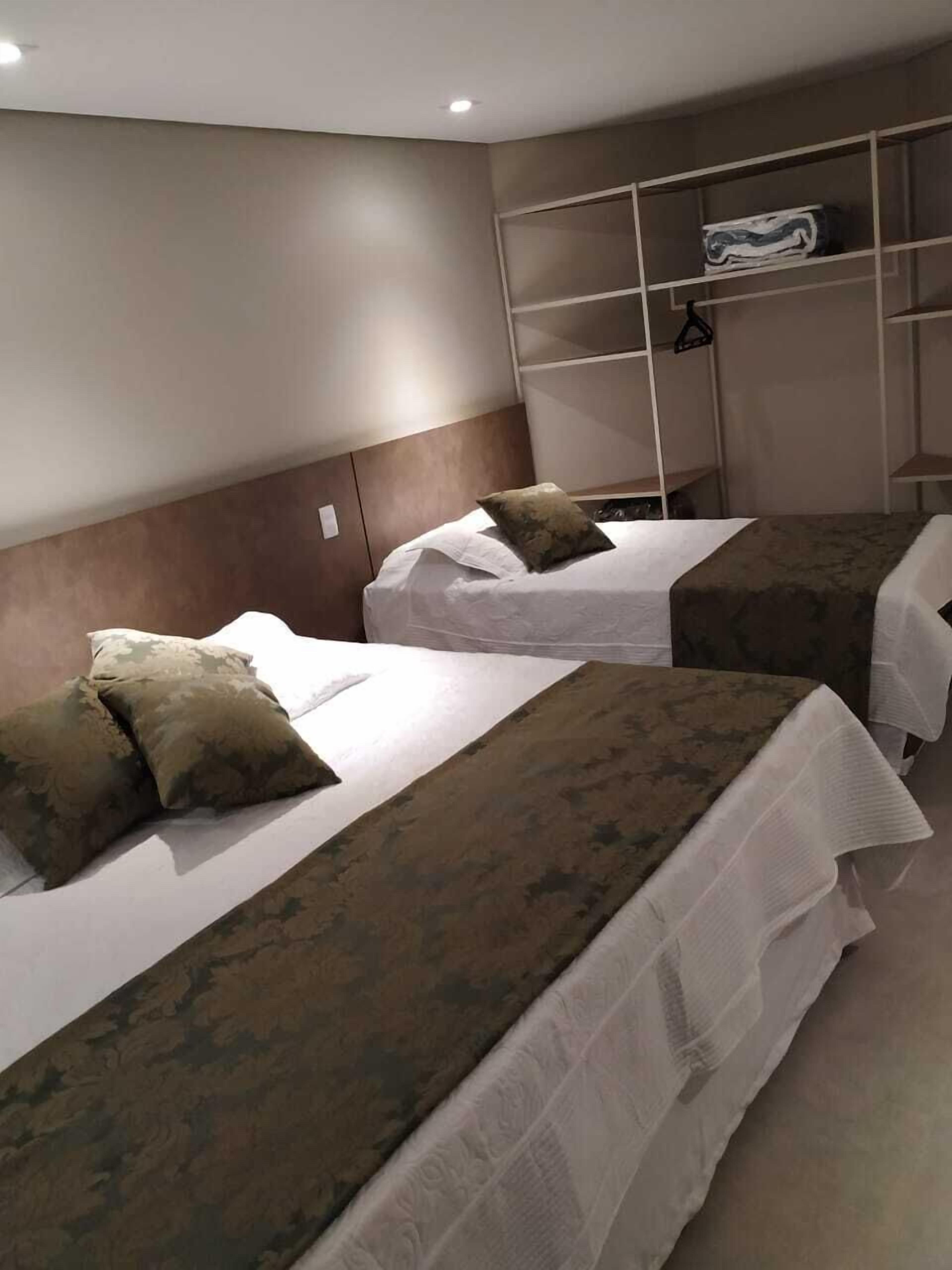 Habitación de hotel con dos camas, cada una con cubrecama blanco y manta marrón. Se observa un armario con estantes y barra para colgar.