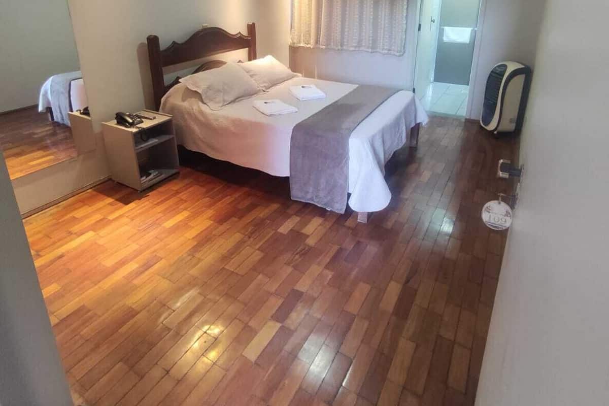 Ein Schlafzimmer mit Holzboden, einem Bett mit weißen Leinen, einem Nachttisch, einem Deckenventilator und einem Fenster mit Vorhängen.