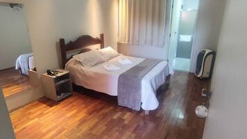 A bedroom with a wooden floor, a bed with white linens, a nightstand, a ceiling fan, and a window with curtains.