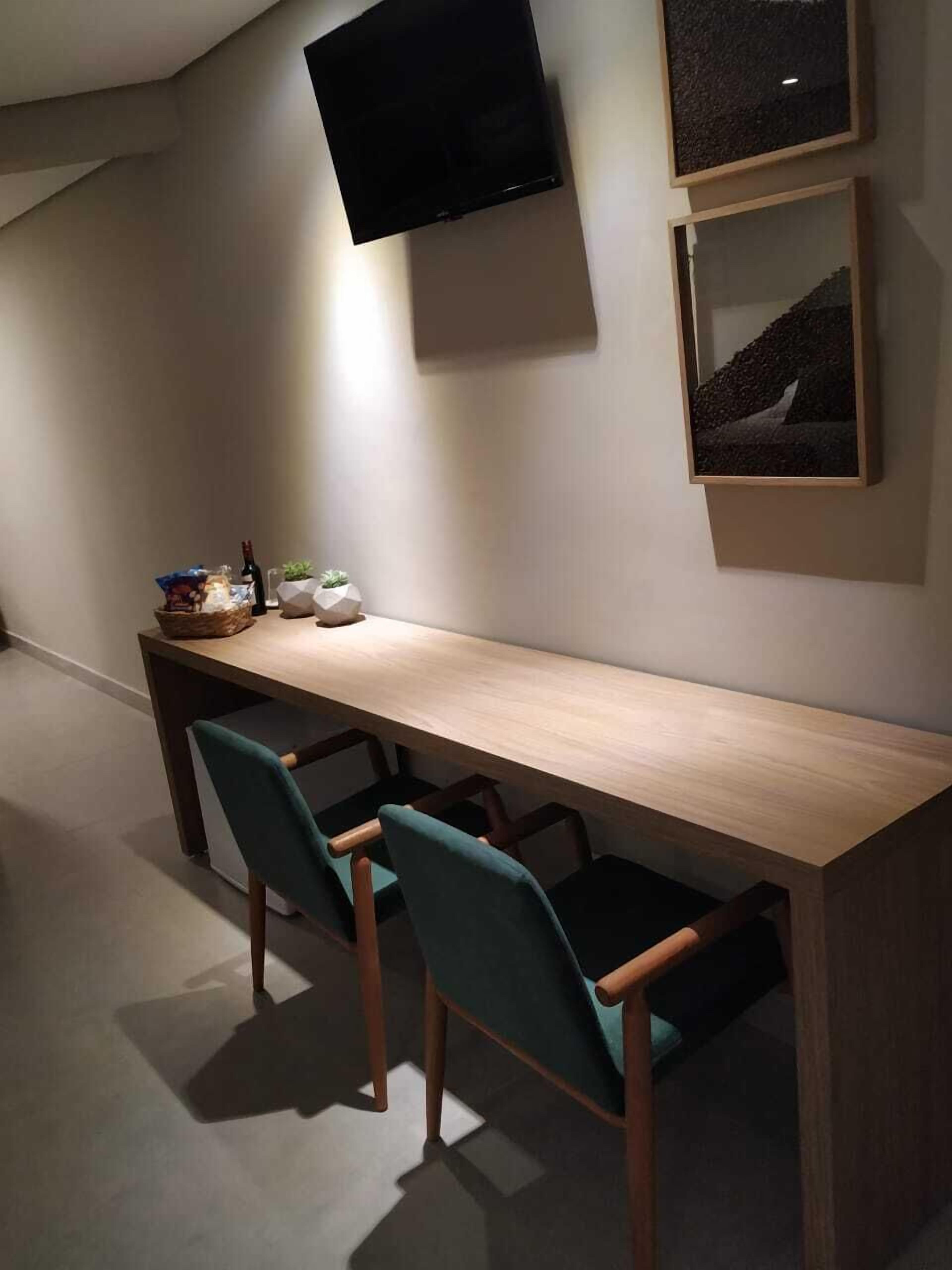 Een moderne kamer met een lang houten bureau, twee blauwgroene stoelen, een flatscreen televisie aan de muur en een kleine potplant op het bureau.