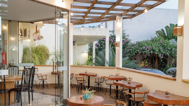 Un café en plein air avec des tables rondes en bois et des chaises en métal, entouré de murs de verre et de grandes fenêtres donnant sur un jardin où s’épanouissent des fleurs roses et des plantes vertes.