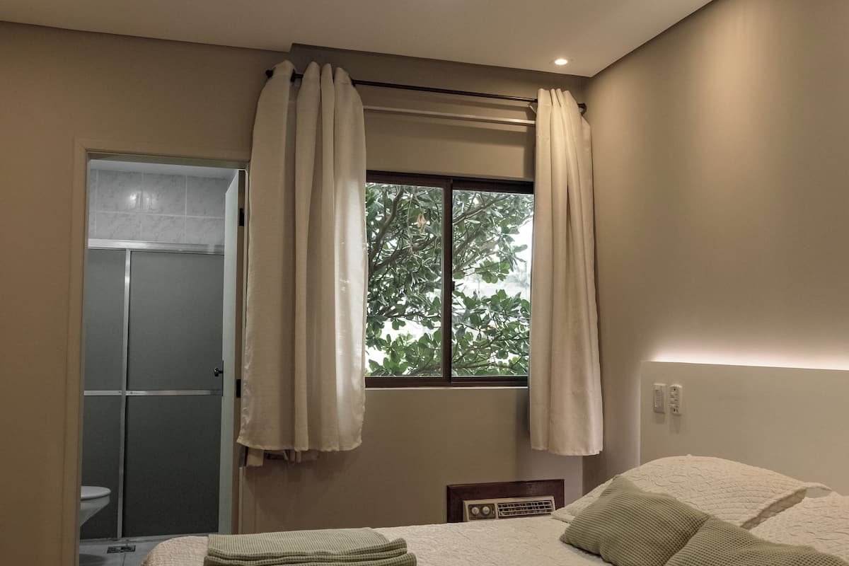 Ein Schlafzimmer mit einem Bett, einem Deckenventilator, einem Fenster mit Vorhängen, einem Nachttisch und einem Beistelltisch.