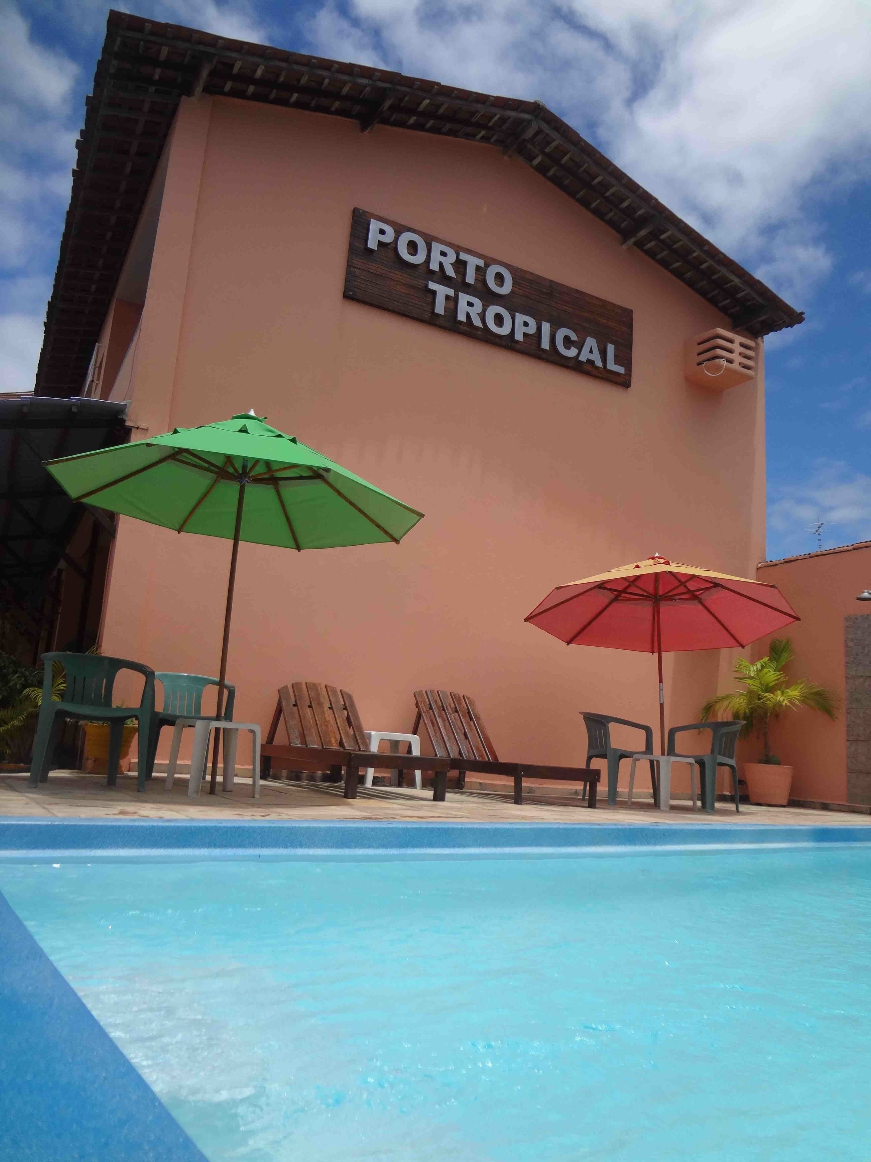 A pool area with lounge chairs and umbrellas, a sign reading "PORTO TROPICAL", and a clear blue sky.