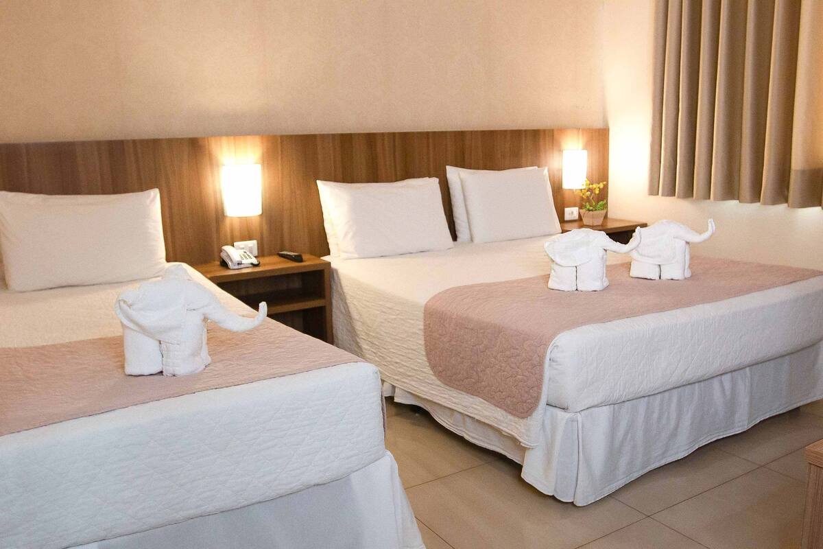Ein Hotelzimmer mit zwei Betten, jeweils mit weißer Decke und einem in Elefant geformten Handtuch. An den Nachttischen stehen Lampen und ein Telefon.