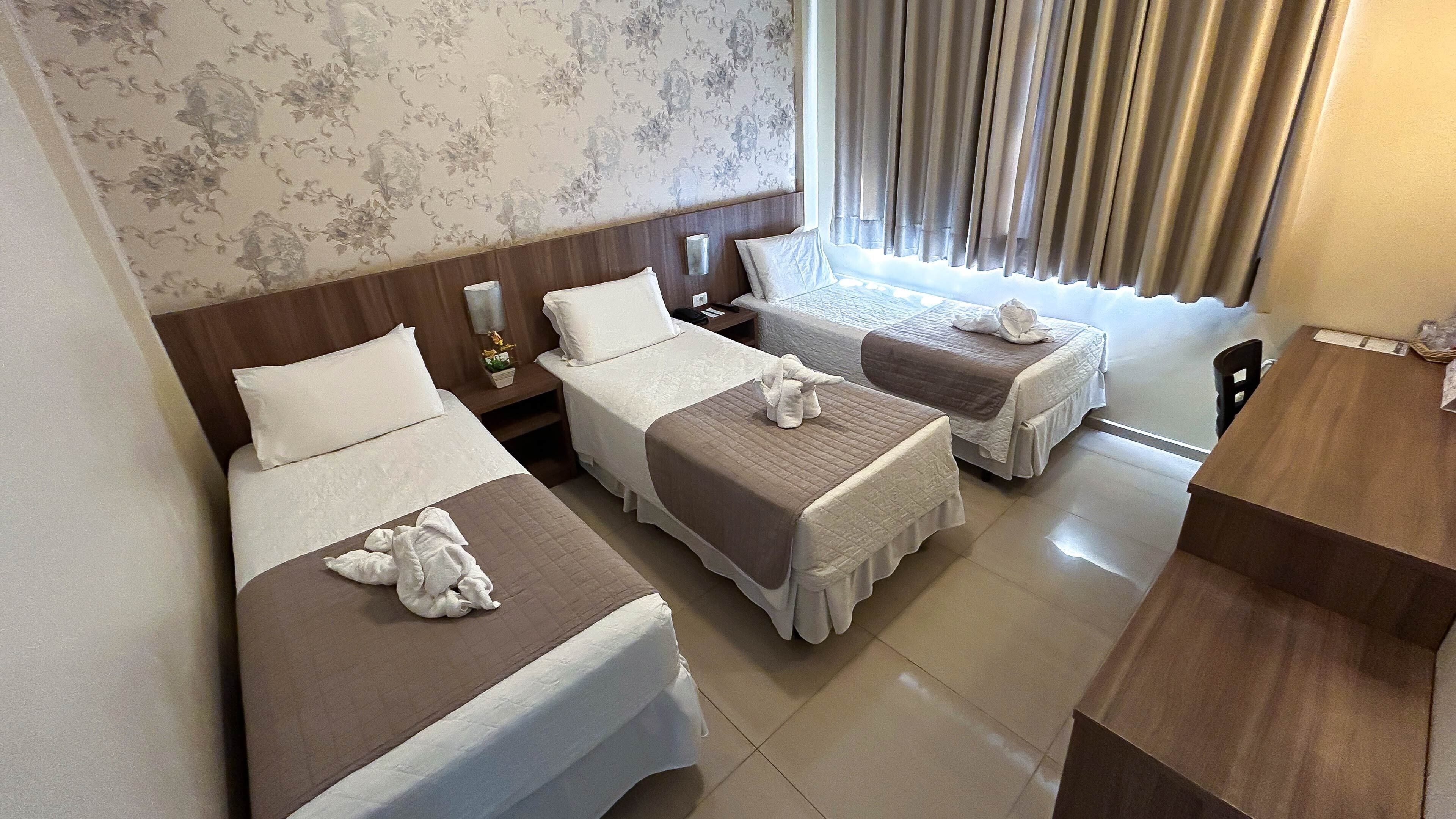 A hotel room with three beds, each with a white and brown bedspread, and a wooden headboard. There is a wooden table and a small nightstand with a lamp.
