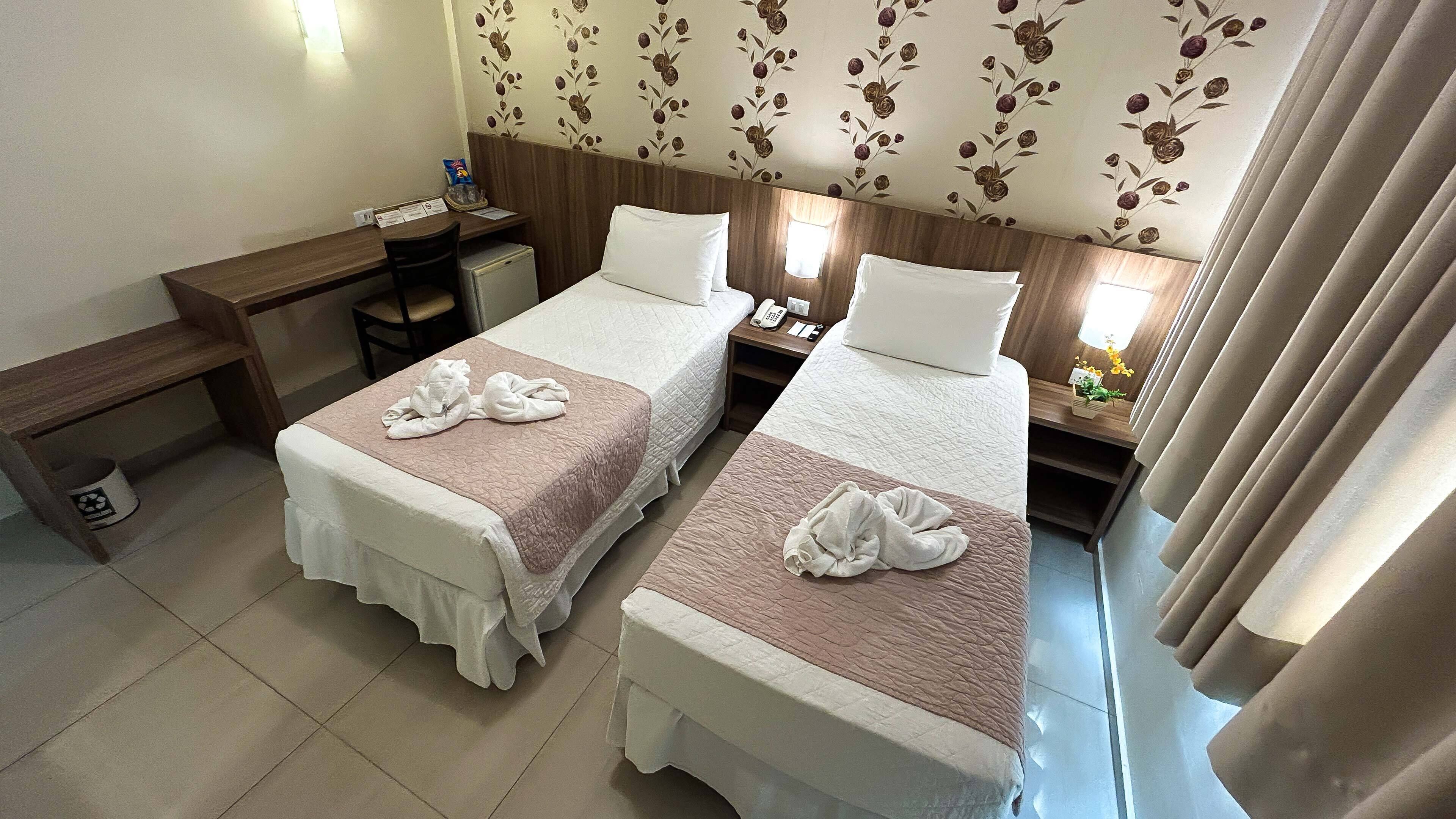 A hotel room with two beds, a desk, a chair, a small table, and a nightstand with a phone and a lamp.