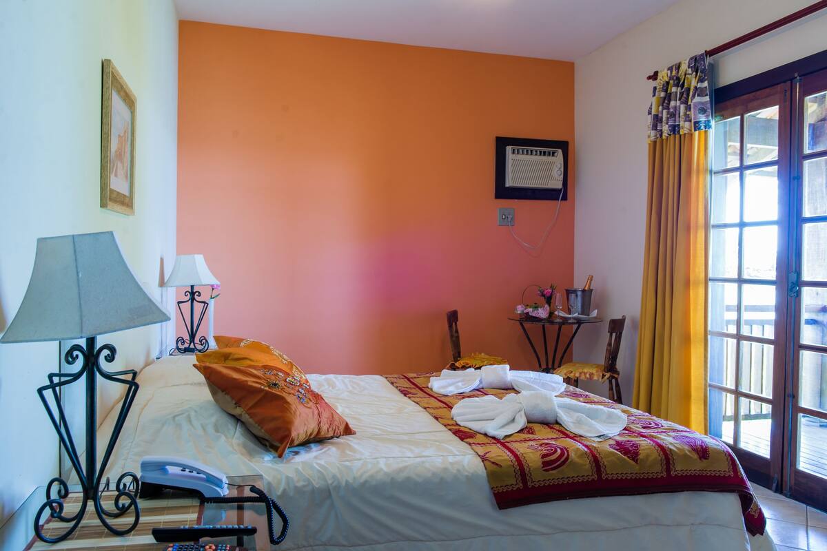 A hotel room with an orange wall, a bed with white bedding, a lamp, a telephone, and a window with curtains.