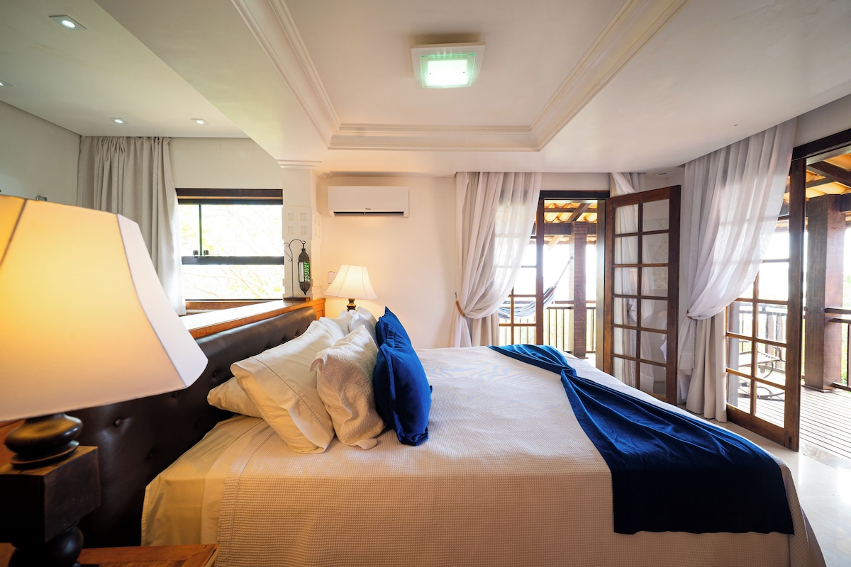 A hotel room with a large bed, a nightstand with a lamp, a window with curtains, and a view of the outdoors.
