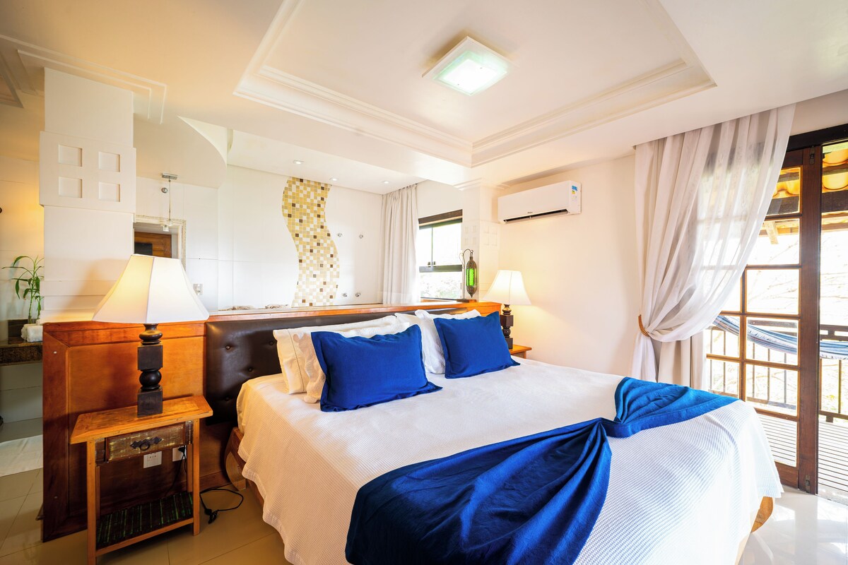 A hotel room with a bed, two bedside tables with lamps, a mirror, a window with curtains, and an air conditioning unit.