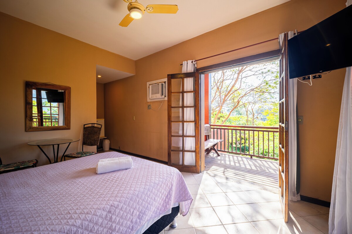 A hotel room with a bed, a small table, a chair, a ceiling fan, a window, and a balcony with a view of trees.