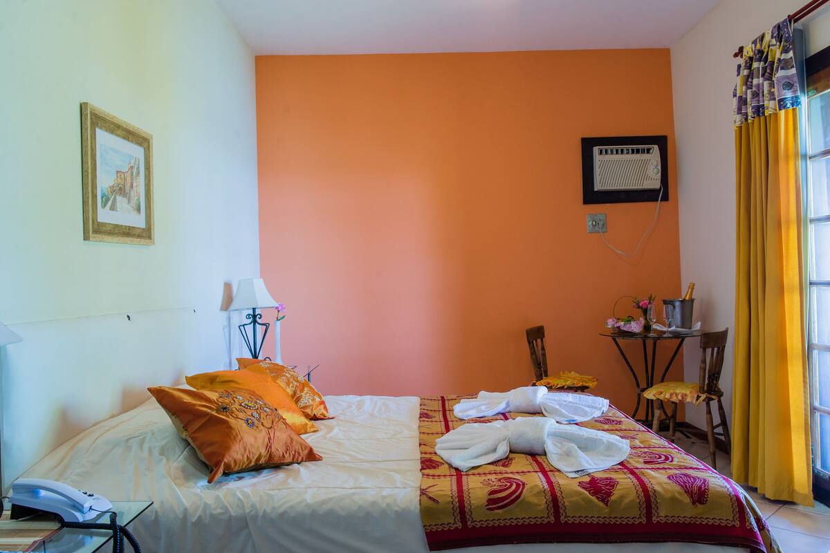 A hotel room with a bed, a bedside table with a lamp, a chair, a small table with a radio, and a window with curtains.