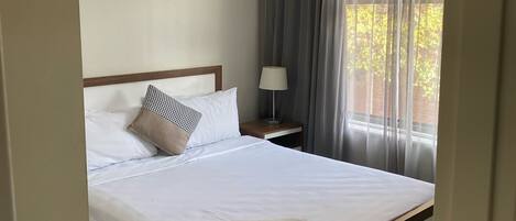 Una cama bien hecha con sábanas blancas y una toalla doblada, una lámpara de noche y una ventana con cortinas.