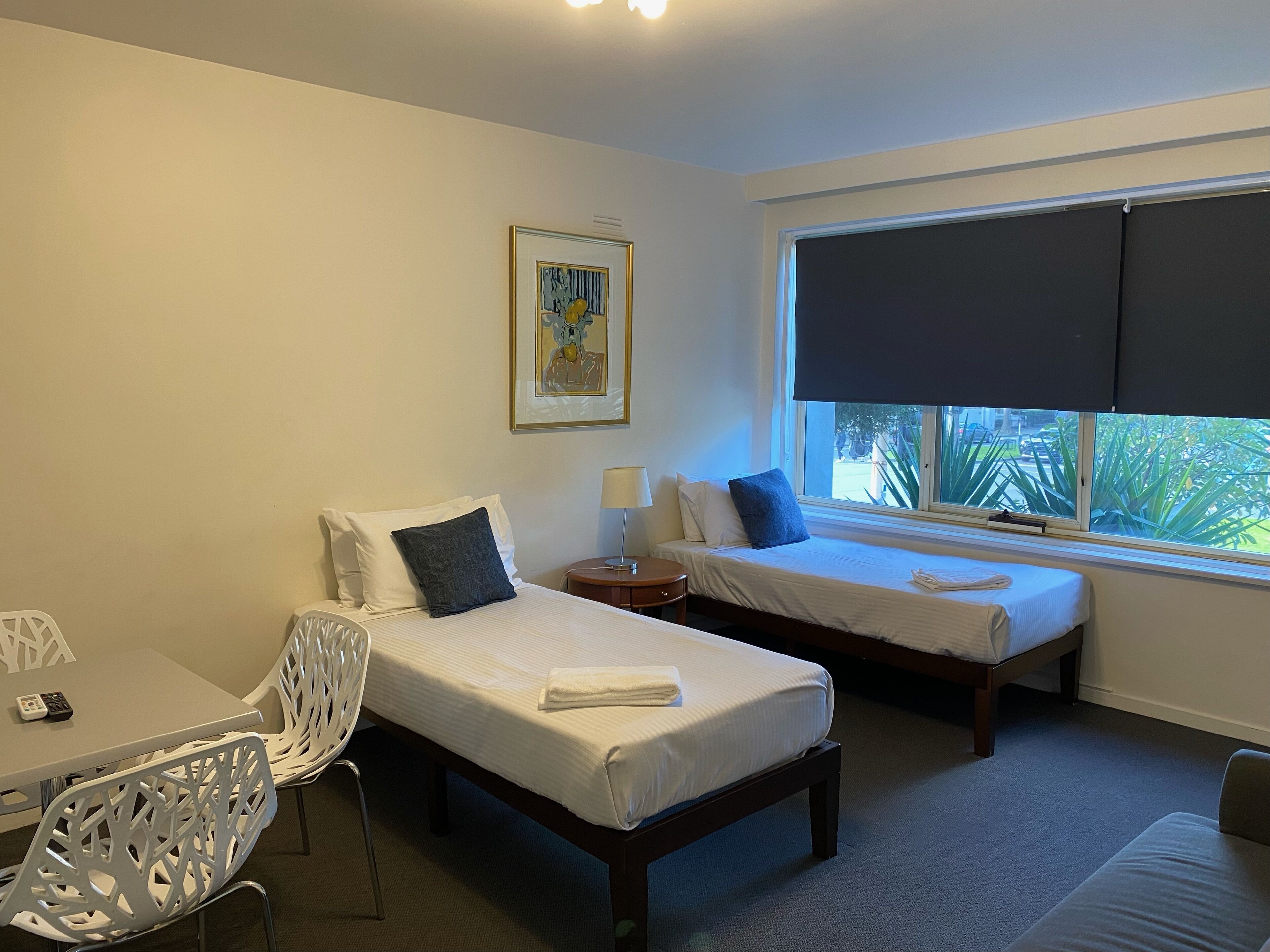 A hotel room with two single beds, a desk with a chair, a window with a view of the outdoors, and a painting on the wall.
