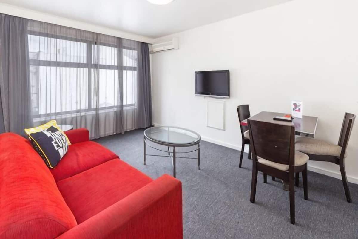 Una habitación de hotel moderna con un sofá rojo, una mesa de centro de vidrio, una mesa de comedor con sillas, un televisor de pantalla plana y una ventana con cortinas.