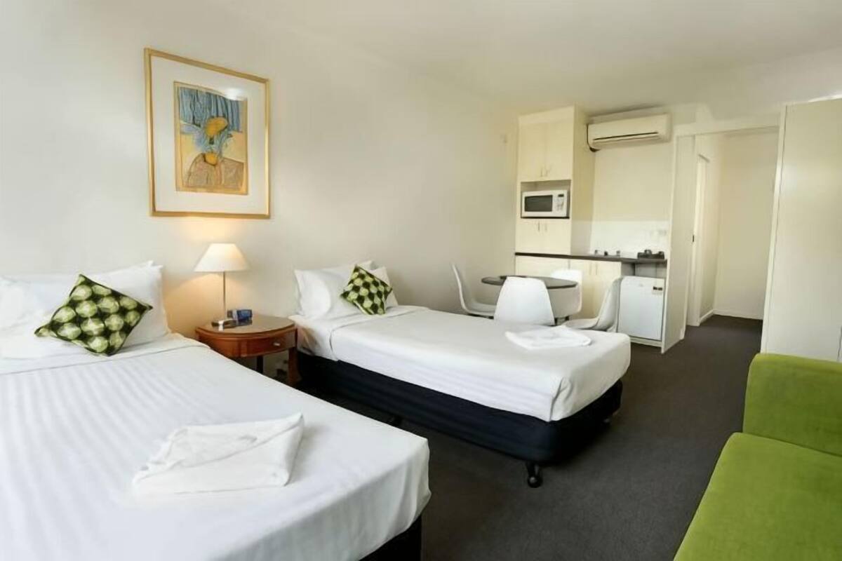 Una habitación de hotel con dos camas, un sillón verde, un escritorio con silla, un microondas y una mesita de noche con lámpara.