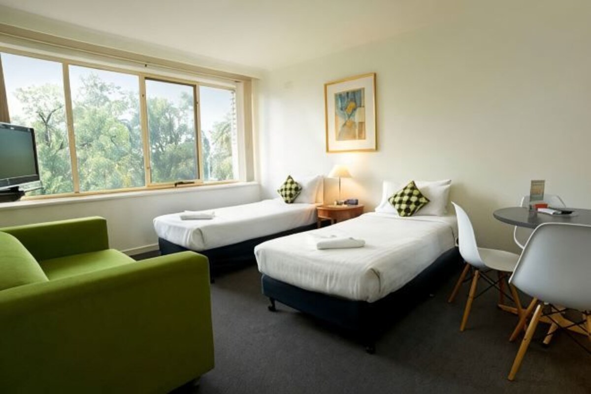 Habitación de hotel con dos camas, una televisión, un escritorio con silla y una ventana que ofrece vista a árboles.