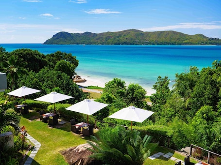 A coastal view with a beach, lush greenery, and outdoor seating with umbrellas.