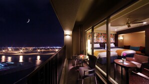 Ein Hotelbalkon mit Blick auf eine nächtliche Stadtlandschaft und einen Fluss. Auf dem Balkon befinden sich zwei Betten, ein Tisch und Stühle.