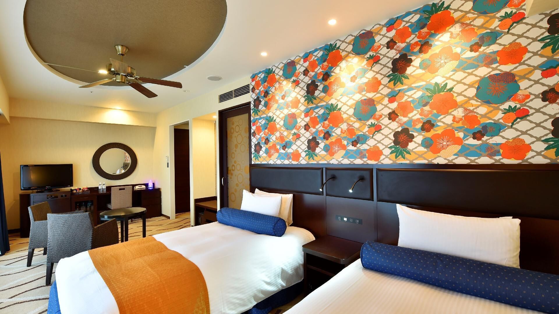 A hotel room with two beds, a desk, a television, and a ceiling fan.