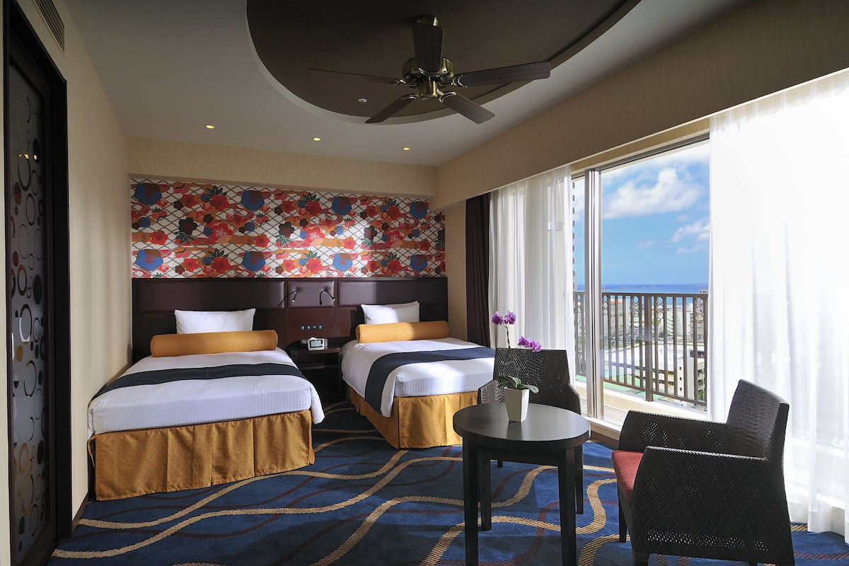 Ein Hotelzimmer mit zwei Betten, einem Deckenventilator, einem großen Fenster mit Blick und einem kleinen Tisch mit Stuhl.
