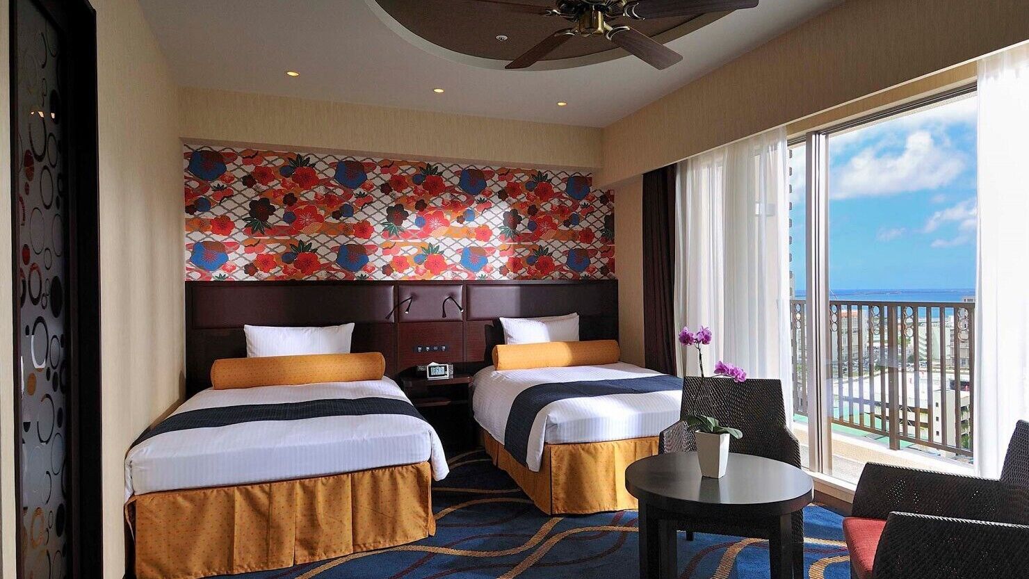 A hotel room with two beds, a ceiling fan, a large window with a view, and a decorative wall.
