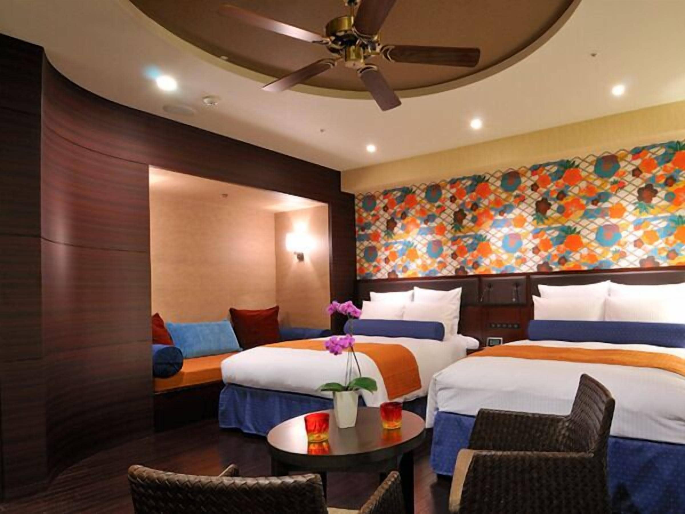 A hotel room with two beds, a ceiling fan, a small table with a vase, and a colorful wall mural.