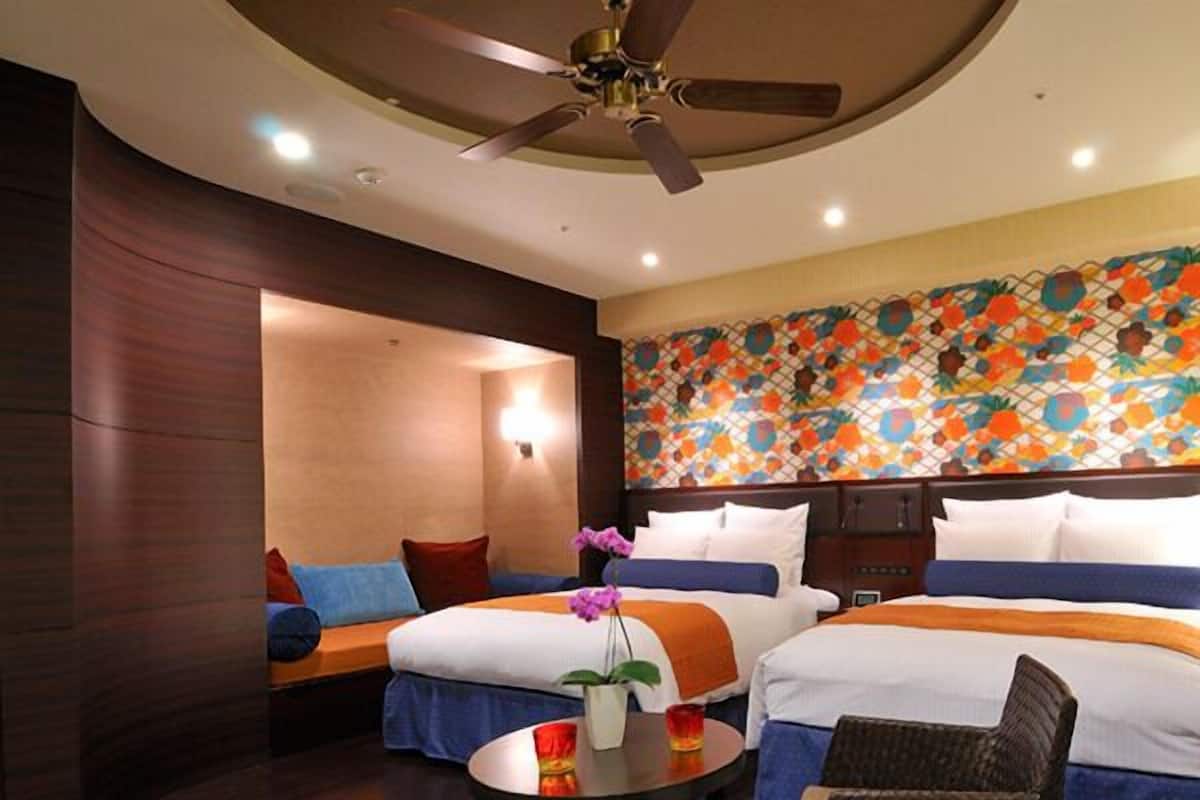 Ein Hotelzimmer mit zwei Betten, einem Deckenventilator, einem kleinen Tisch mit einer Vase und einem farbenfrohen Wandbild.