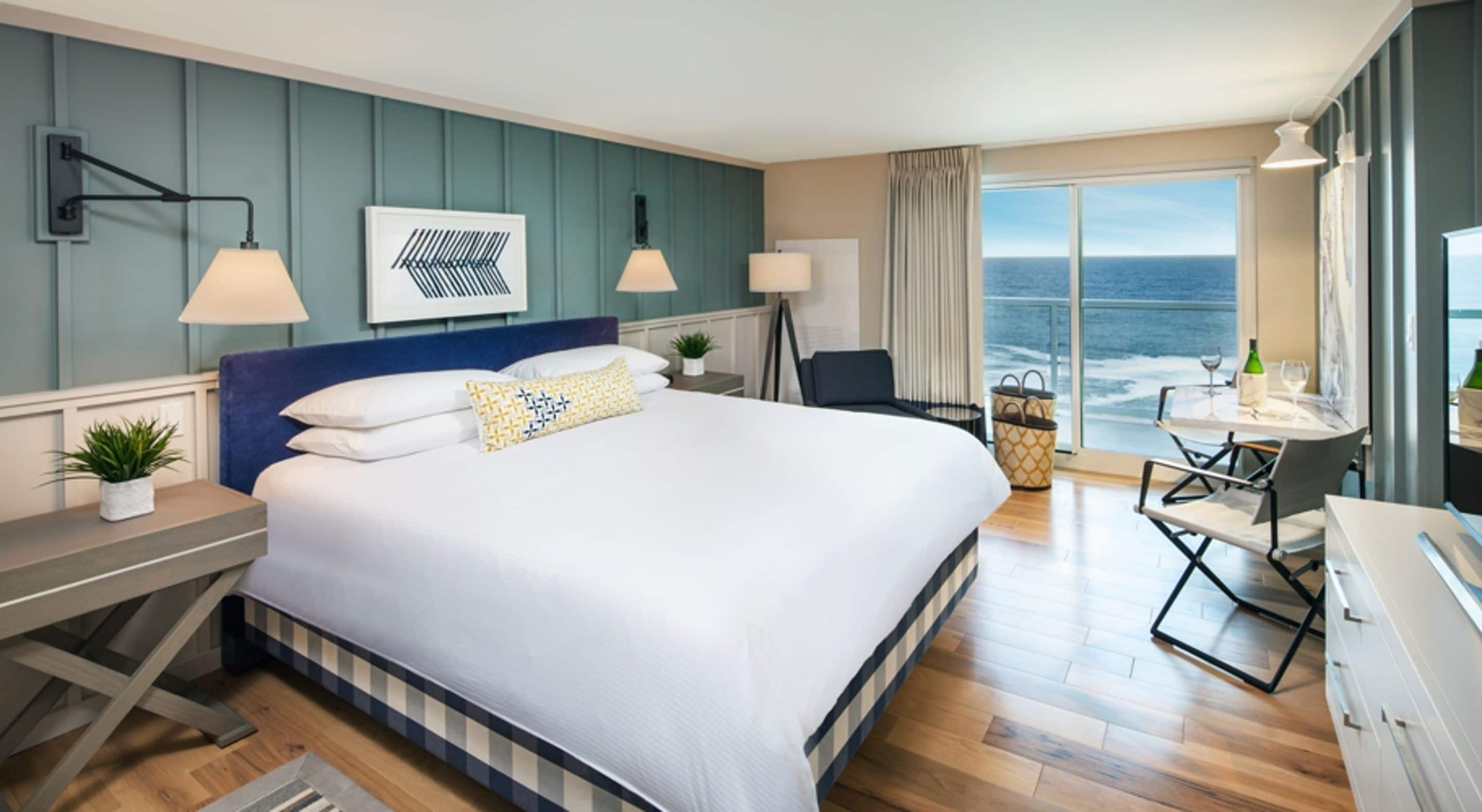 A hotel room with a large bed, a desk with a chair, a view of the ocean, and a dining area with a table and chairs.