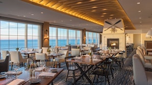 A restaurant with tables set for dining, large windows offering ocean views, and a fireplace. - Cliff House Maine (Cape Neddick)