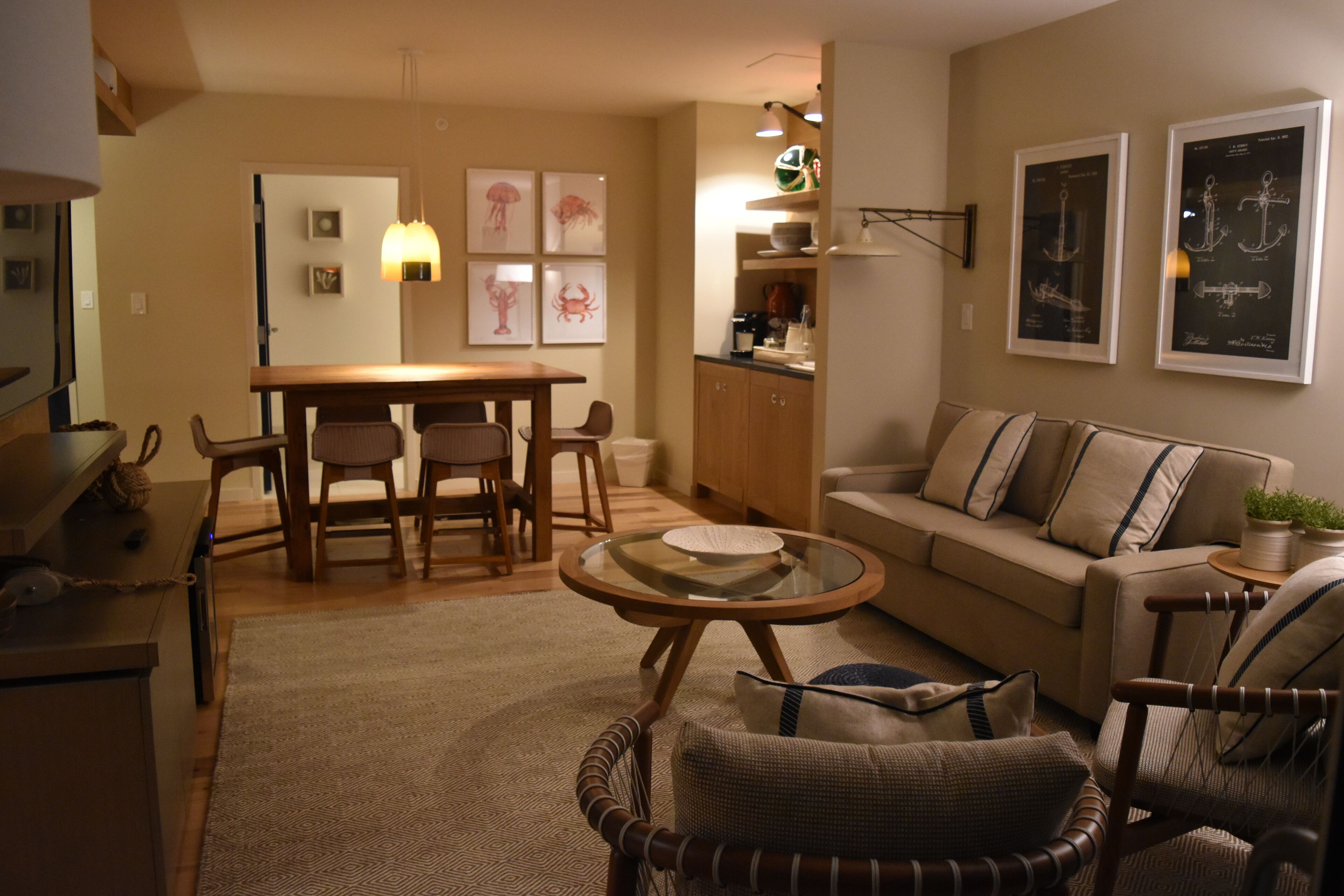 A cozy living room with a wooden dining table, a glass-top coffee table, a sofa, and a wicker armchair. There are framed pictures on the wall and a kitchen area in the background.