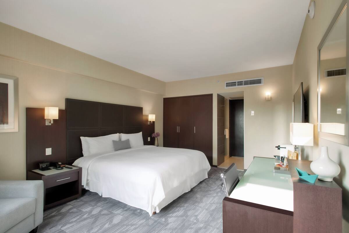 A hotel room with a large bed, a bedside table with a lamp, a desk with a mirror, and a bathroom door.