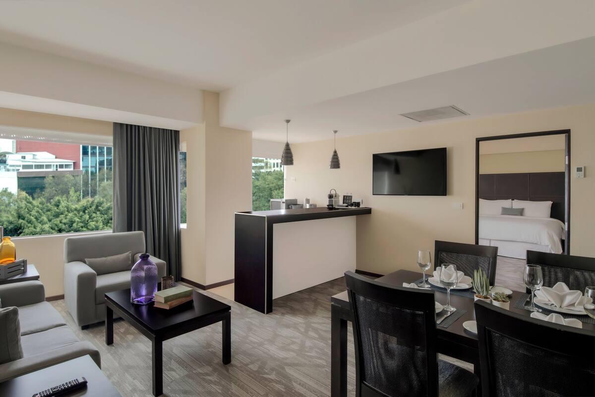 A hotel room with a sofa, a dining table set for two, a kitchen area, a large bed, and a TV mounted on the wall.