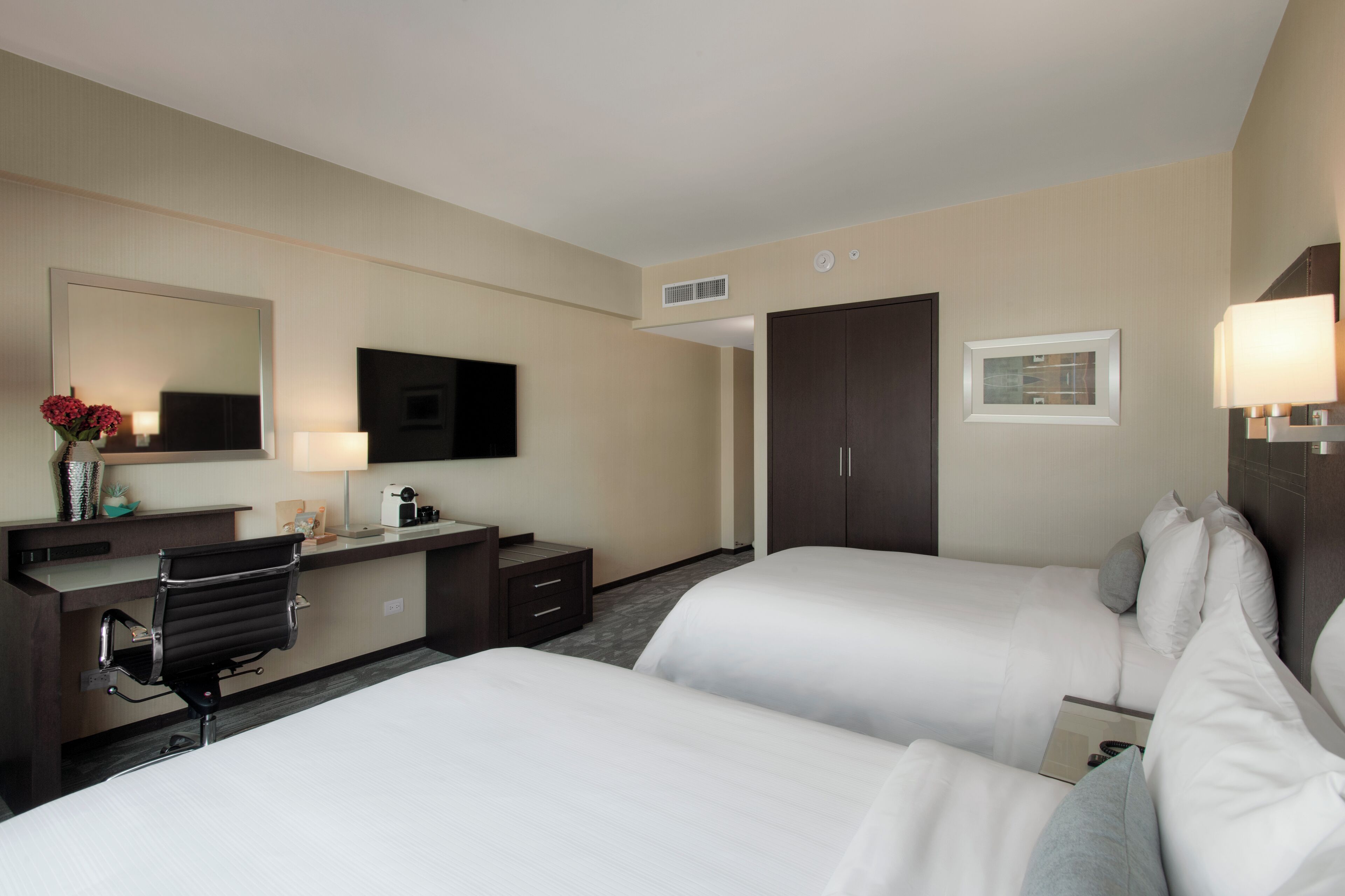 A hotel room with two beds, a desk with a chair, a flat-screen TV, a mirror, and a door.