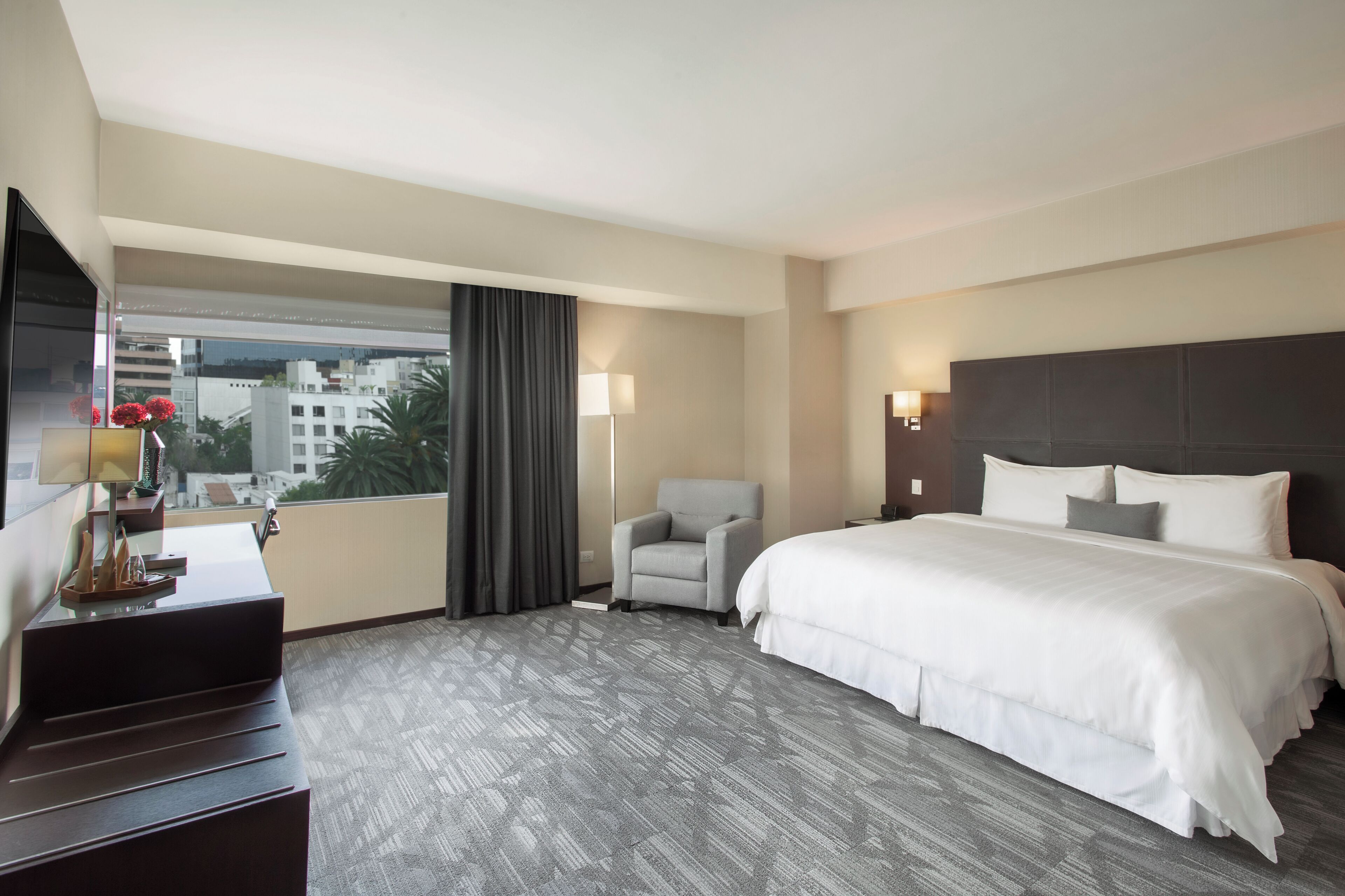 Habitación de hotel con una cama grande, un escritorio con lámpara, una silla y vistas a edificios a través de una ventana.