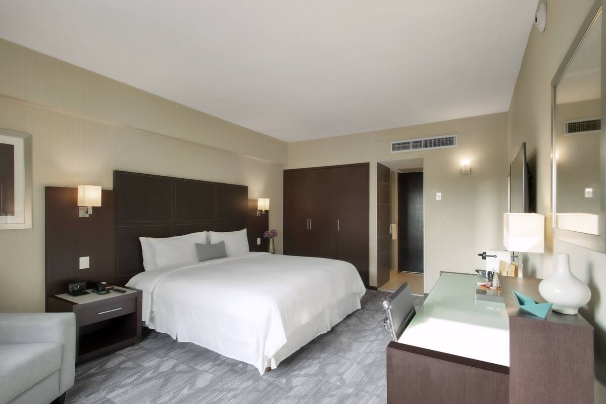 A hotel room with a large bed, a bedside table with a lamp, a desk with a mirror, and a bathroom door.