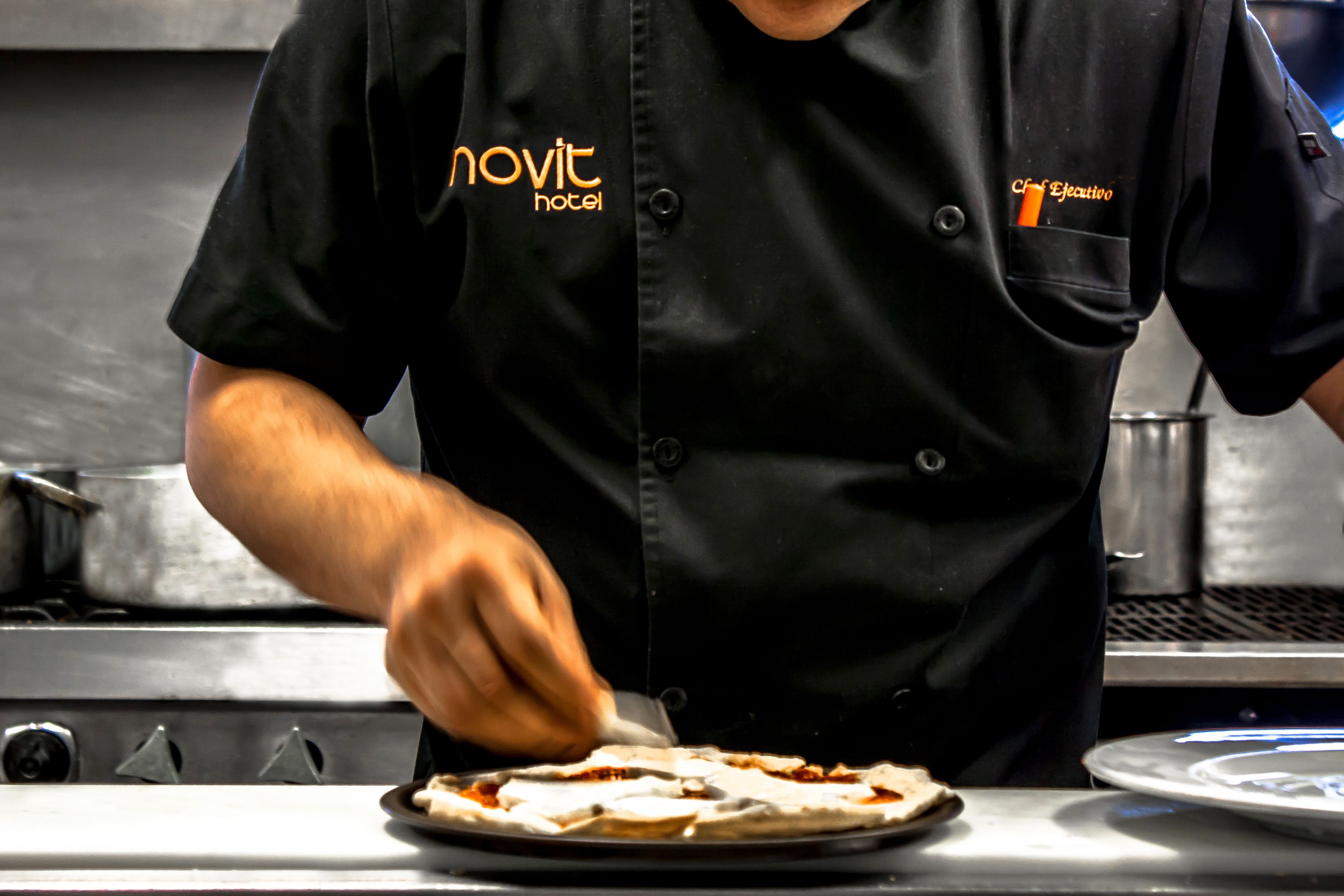 Un chef vêtu d’un uniforme noir, orné des inscriptions « Novit Hotel » et « Chef Ejecutivo », prépare des plats dans une cuisine.