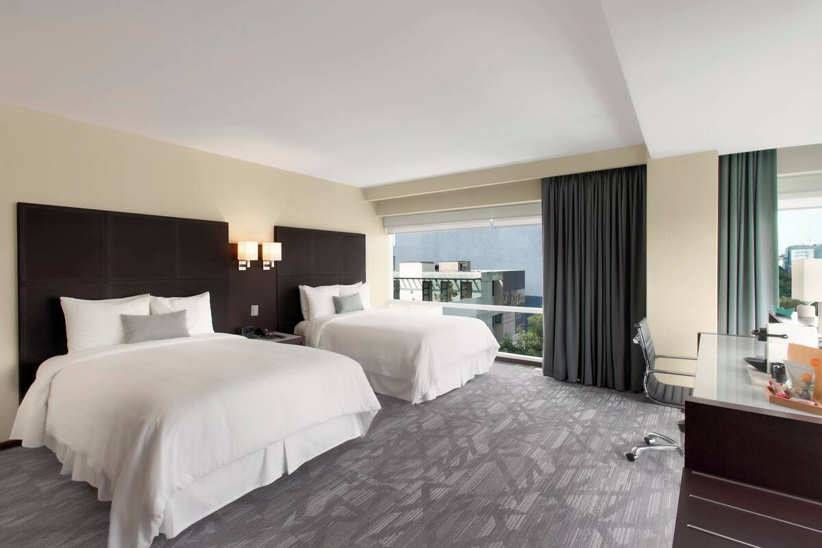 A hotel room with two beds, a desk, and a large window with a view of the city.