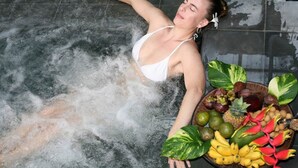 Una persona con traje de baño blanco recostada en un jacuzzi, con un cuenco de frutas tropicales cerca.