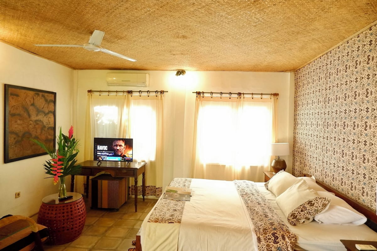 A hotel room with a bed, a desk with a television, a red decorative vase, and patterned wallpaper.