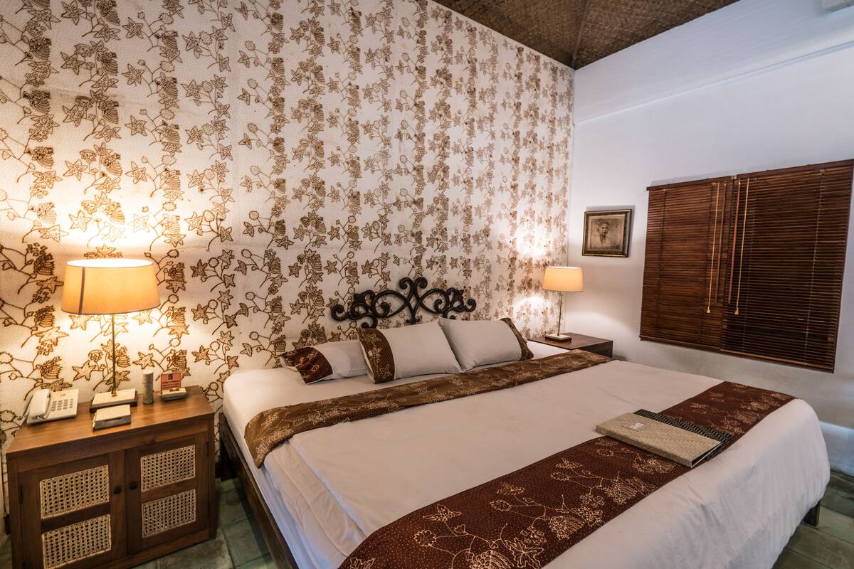 A hotel room with a large bed, floral wallpaper, wooden headboard, bedside lamps, and a window with blinds.