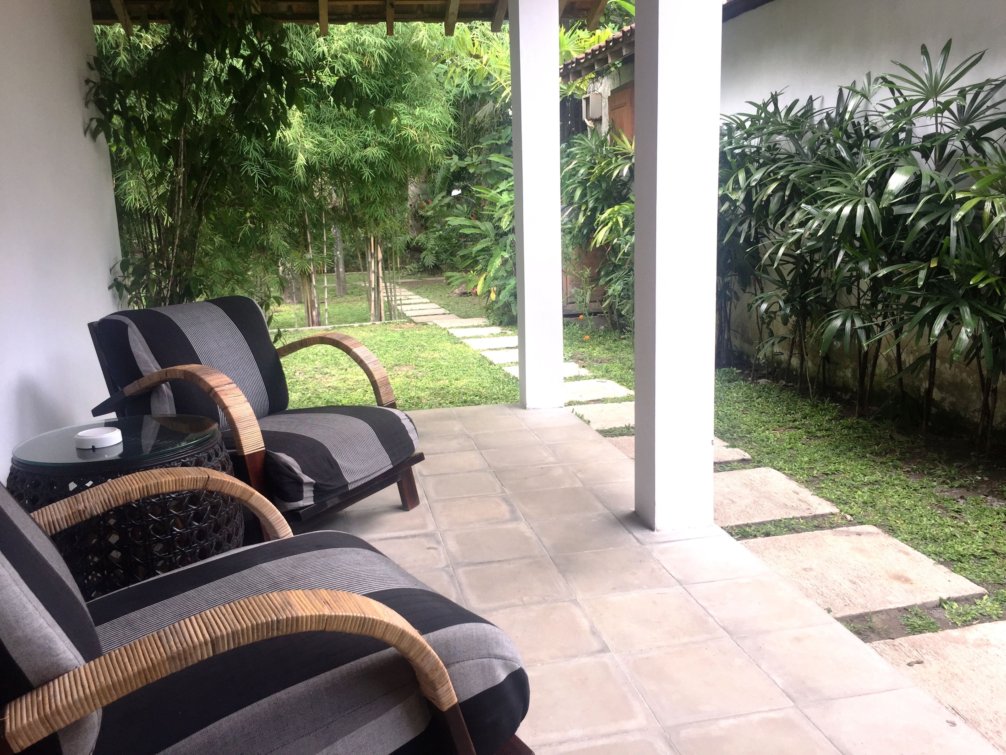 A covered outdoor seating area with two wicker chairs and a glass table, surrounded by lush greenery and a stone pathway.
