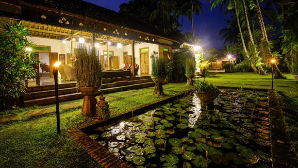 Un edificio tradicional con un porche cubierto, un pequeño estanque con nenúfares y un jardín bien cuidado con palmeras.