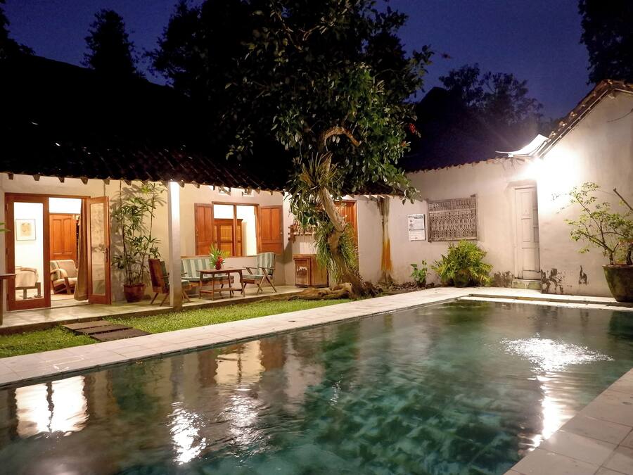 A swimming pool with a tiled deck, surrounded by a small garden and a building with a tiled roof.