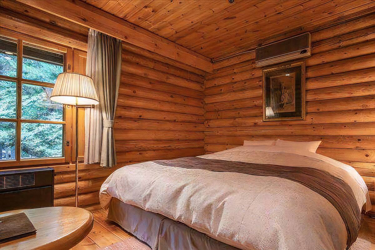 A log cabin bedroom with a large bed, a bedside table, a lamp, and a window with curtains.